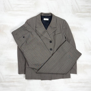 May include: A grey herringbone patterned blazer and matching trousers set. The blazer features a notched lapel, two dark buttons, and a dark navy interior lining. The trousers have a straight-leg cut with a single visible button on the pocket.