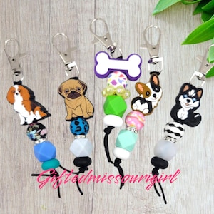 May include: Five colorful keychains with different dog breeds, including a Cavalier King Charles Spaniel, a Pug, a Corgi, and a Husky. Each keychain features a black cord with colorful beads and a silver clip.