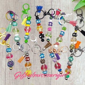 May include: A collection of colorful beaded keychains with various charms and tassels. Each keychain features a different combination of beads, including clear, patterned, and solid colors. The keychains are attached to silver key rings.
