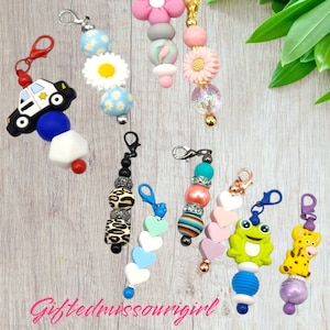 May include: Assortment of colorful beaded keychains with various charms, including a police car, a frog, a leopard print bead, a flower, and a giraffe. The keychains are made with different types of beads, including plastic, wood, and metal.