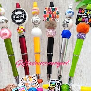 May include: A collection of decorative pens with various designs. The pens feature colorful beads, pom-poms, and text that reads "Social Worker" and "Work". The pens have different colored barrels, including green, red, yellow, black, and silver.