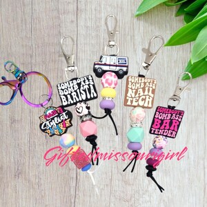 May include: A collection of keychains with various charms and beaded accents. Charms include a postal truck, and text that reads "Somebody's Bomb Ass Barista", "Nail Tech", "Bar Tender", and "Hair Stylist". The keychains have silver-tone clasps.