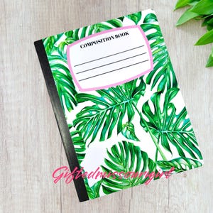 May include: Composition book with a tropical leaf design. The cover features large green monstera leaves on a white background. The top has a pink-bordered label that reads "COMPOSITION BOOK". The spine is black.