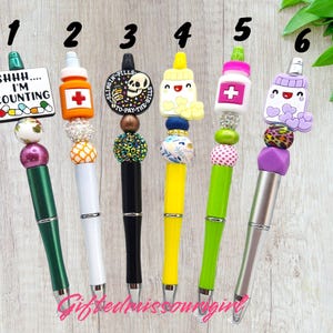 K&ouml;nnte beinhalten: Sechs dekorative Stifte mit einzigartigen Designs. Jeder Stift hat einen anderen Aufsatz, darunter eine Pillenflasche, ein Totenkopf und ein Schild mit der Aufschrift "Shhh... I'm counting." Die Stifte haben bunte Perlen und Akzente.