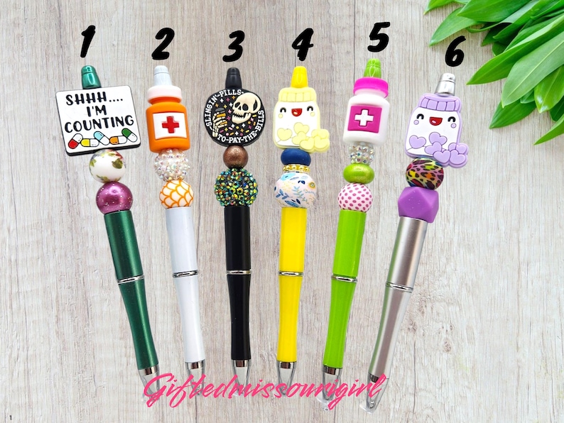 K&ouml;nnte beinhalten: Sechs dekorative Stifte mit einzigartigen Designs. Jeder Stift hat einen anderen Aufsatz, darunter eine Pillenflasche, ein Totenkopf und ein Schild mit der Aufschrift "Shhh... I'm counting." Die Stifte haben bunte Perlen und Akzente.