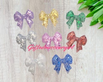 May include: A collection of glittery resin bows in various colors, including purple, gold, green, silver, pink, coral, white, and blue. The bows are arranged on a light wood-grain surface. The bows are decorated with glitter.
