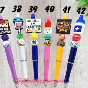 K&ouml;nnte beinhalten: Eine Sammlung von Stiften mit Perlen und verschiedenen Designs. Jeder Stift hat einen einzigartigen Aufsatz, darunter eine Pille, eine Spritze und ein Schild zum Thema Apotheke. Die Stifte sind in einer Vielzahl von Farben erh&auml;ltlich, wie z. B. Lila, Wei&szlig;, Blau, Rosa und Gelb.