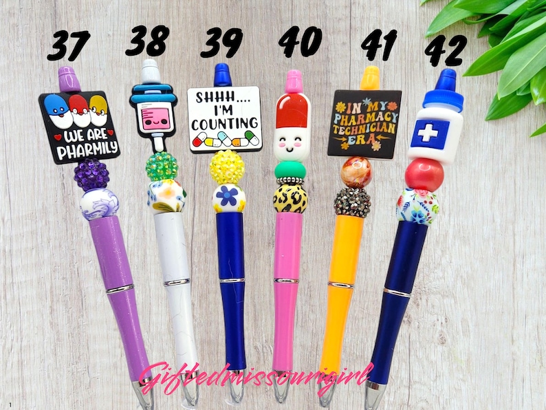 K&ouml;nnte beinhalten: Eine Sammlung von Stiften mit Perlen und verschiedenen Designs. Jeder Stift hat einen einzigartigen Aufsatz, darunter eine Pille, eine Spritze und ein Schild zum Thema Apotheke. Die Stifte sind in einer Vielzahl von Farben erh&auml;ltlich, wie z. B. Lila, Wei&szlig;, Blau, Rosa und Gelb.