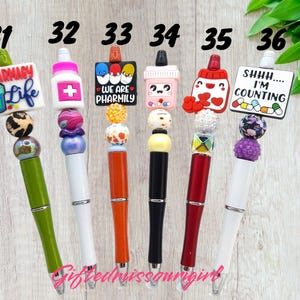 K&ouml;nnte beinhalten: Sechs bunte Stifte mit dekorativen Aufs&auml;tzen. Die Stifte haben verschiedene Designs, darunter Apotheken-Themen-Anh&auml;nger mit Texten wie "Pharmacy Life" und "Shhh... I'm Counting." Jeder Stift hat ein einzigartiges Farbschema und Perlenakzente.