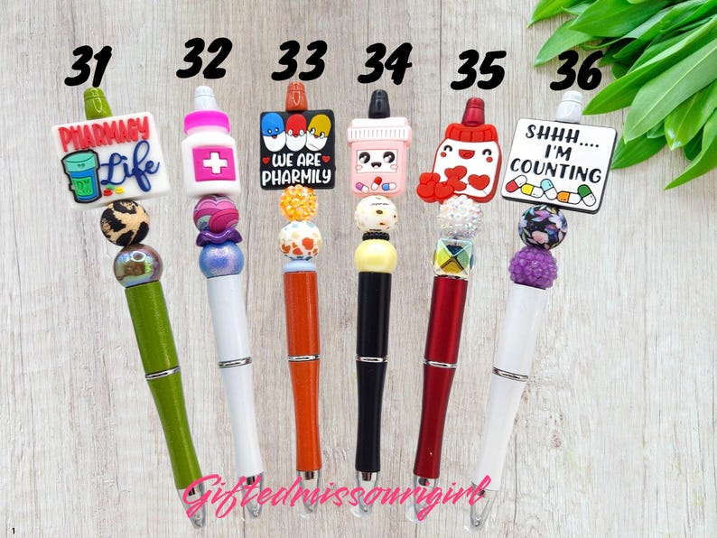 K&ouml;nnte beinhalten: Sechs bunte Stifte mit dekorativen Aufs&auml;tzen. Die Stifte haben verschiedene Designs, darunter Apotheken-Themen-Anh&auml;nger mit Texten wie "Pharmacy Life" und "Shhh... I'm Counting." Jeder Stift hat ein einzigartiges Farbschema und Perlenakzente.
