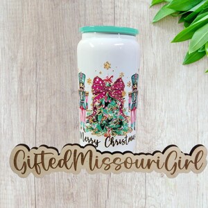 May include: White tumbler with a teal rim featuring a Christmas tree design, pink bow, and nutcracker soldiers. The words "Merry Christmas" are printed below the tree. The tumbler is cylindrical and stands on a wooden surface.