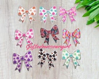 May include: Nine decorative bow-shaped embellishments with floral patterns in various colors, including pink, orange, blue, and purple. The bows are arranged on a light wood-grain surface. The text "Giftedmissourigirl" is visible.