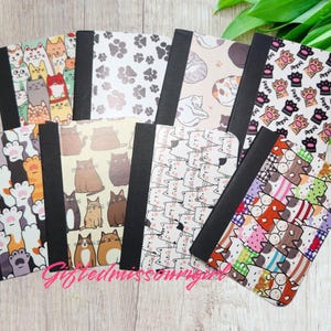 May include: A collection of notebooks with various cat-themed designs. The covers feature colorful illustrations of cats, paw prints, and patterns. Each notebook has a black spine and a variety of colors, including orange, brown, and white.
