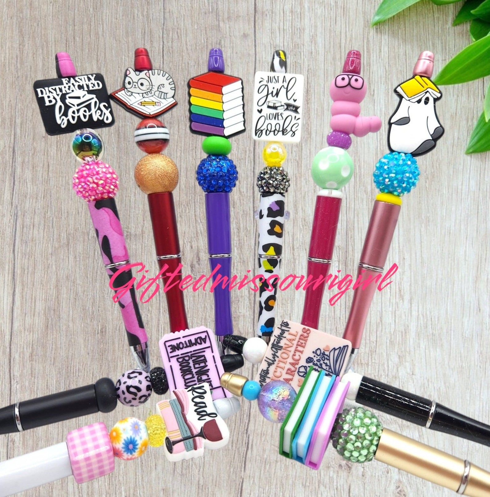 A light wooden background with a bunch of colorful pens on it, with large charms like rainbow books, a "bookworm", a ghost with a book, and colorful beads.