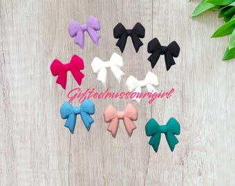 May include: A collection of colorful bow-shaped accessories in various colors, including purple, black, white, pink, blue, and teal. The bows are arranged on a light wood-grain surface. The text "Giftedmissourigirl" is visible.