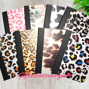 May include: A collection of notebooks with various animal print covers, including leopard, cow, and cheetah patterns. The notebooks have black spines and are arranged in a fan shape. The text "Giftedmissouri" is visible in the image.