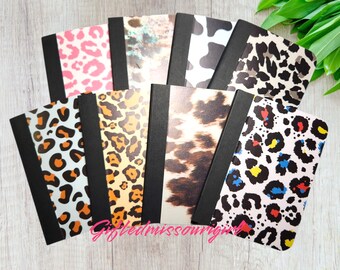 May include: A collection of notebooks with various animal print covers, including leopard, cow, and cheetah patterns. The notebooks have black spines and are arranged in a fan shape. The text "Giftedmissouri" is visible in the image.