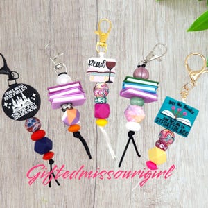 May include: Five colorful book-themed keychain charms with different sayings. The charms feature a variety of beads and charms, including books, wine glasses, and flowers. The charms are attached to black cords with silver clasps.