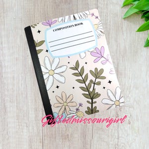 May include: A composition book with a floral design featuring daisies and other flowers in white, lavender, and tan. The cover has the words "COMPOSITION BOOK" and lines for a name. The notebook is on a light wood surface.
