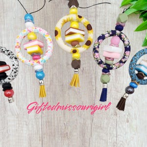 May include: Five colorful car charms with various designs. Each charm features a ring with decorative elements, including beads, tassels, and miniature objects resembling books and desserts. The charms are suspended from black cords.