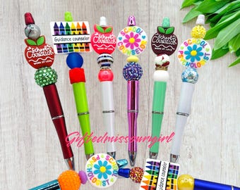 May include: A collection of decorative pens with various toppers. Some pens feature apple designs with "School Counselor" text, while others have flower or crayon designs. The pens come in a variety of colors, including red, green, white, and purple.