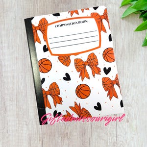 May include: Composition book with a white cover featuring orange basketballs, bows, and black hearts. The cover has the words "COMPOSITION BOOK" and lines for writing. The notebook has a black spine.