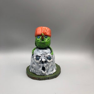 May include: A green and orange ceramic sculpture of a small figure with a large head wearing a red hat sitting on top of a larger white ceramic head with blue eyes.