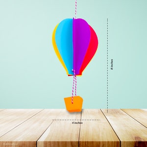 K&ouml;nnte beinhalten: Eine bunte Hei&szlig;luftballon-Dekoration, etwa 20 cm hoch und 10 cm breit, h&auml;ngt an einer Schnur. Der Ballon hat gelbe, blaue, violette und rote Paneele. Ein orangefarbener Korb h&auml;ngt darunter.