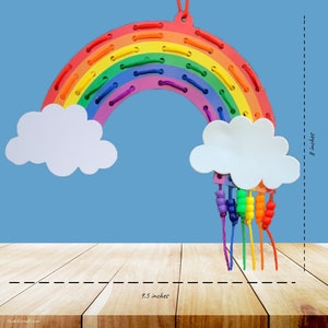 K&ouml;nnte beinhalten: Ein regenbogenf&ouml;rmiges Spielzeug, ca. 24 cm breit und 20 cm hoch, mit einem gen&auml;hten Design und wei&szlig;en Wolkenakzenten. Der Regenbogen hat die Farben Rot, Orange, Gelb, Gr&uuml;n, Blau und Lila. Perlenstr&auml;nge h&auml;ngen von den Wolken.