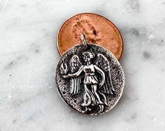 May include: Silver charm pendant featuring a detailed depiction of an angel with wings outstretched. The charm is about the size of a US penny.