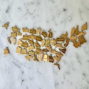 May include: A collection of gold-colored state charms arranged in the shape of the United States. Each charm is a miniature outline of a state, with the state's name engraved on it. The charms are scattered on a white marble surface.