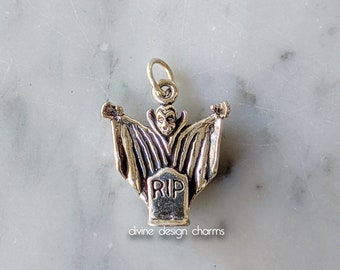 May include: Silver charm featuring a vampire with outstretched wings standing on a tombstone that reads "RIP".