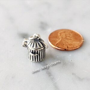 May include: A small, silver-colored trash can charm with a hinged lid and vertical stripes. The charm has a loop for attaching to a bracelet or necklace. The text "divine design charms" is visible. A penny is next to the charm.