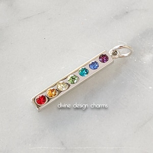 May include: A silver pendant with seven colorful gemstones arranged in a row. The gemstones are red, orange, yellow, green, blue, indigo, and violet. The pendant has a small loop at the top for a chain. The text "divine design charms" is at the bottom.