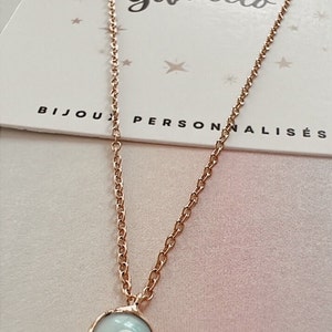 Puede incluir: Un collar de cadena dorada con un colgante azul de ojo turco. El colgante es redondo y tiene un borde blanco. El collar está sobre un fondo rosa.