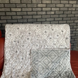 May include: A reversible quilt. One side displays a floral pattern of white flowers on a light grey backdrop. The reverse side is a solid grey with a diamond pattern. The quilt is folded over, revealing both sides.