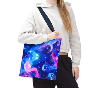 May include: A tote bag with a vibrant, colorful galaxy design. The bag features a deep blue background with swirling clouds of pink, purple, and blue, accented by white stars. The tote bag has a black strap.