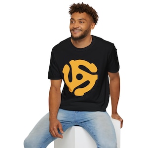 May include: Black t-shirt featuring a large, yellow 45 RPM record adapter graphic on the front. The shirt is made of a soft material and is being worn by a person. The person is wearing blue jeans and is sitting on a white surface.