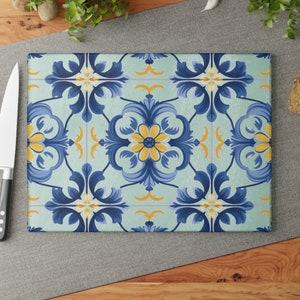 Op de afbeelding: Rechthoekige glazen snijplank met een bloemen tegelpatroon in blauw, geel en wit op een lichtgroene achtergrond. Een roestvrijstalen mes met een zwart handvat bevindt zich aan de linkerkant.