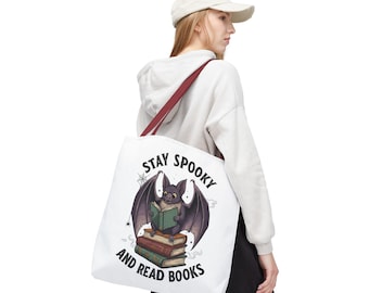 Puede incluir: Una bolsa de tela blanca con una ilustración gráfica de un murciélago leyendo un libro verde mientras está posado sobre una pila de tres libros. El texto "STAY SPOOKY AND READ BOOKS" está impreso en letras mayúsculas negras alrededor.