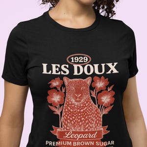 May include: Black t-shirt with a vintage-style design. The design features the text "1929 LES DOUX" above a red and white leopard illustration surrounded by red flowers. Below the leopard is the word "Leopard" on a banner, and the text "PREMIUM BROWN SUGAR" and "LOCALLY GROWN & ETHICALLY HARVESTED".