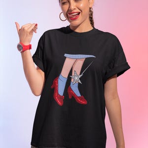Może przedstawiać: Czarny t-shirt z zabawną ilustracją nóg w czerwonych szpilkach, niebieskich skarpetach i spódnicy w kratkę vichy, z błyszczącą gwiazdką. Projekt jest zabawny i przyciąga wzrok.
