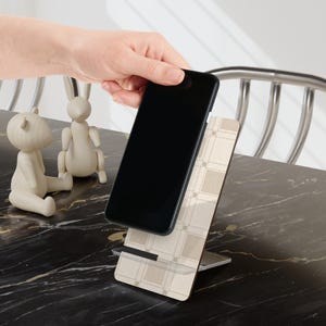 Può includere: Uno smartphone nero è posizionato su un supporto per telefono con motivo beige e bianco. Il supporto ha una base rettangolare e un supporto verticale con un design a griglia. Due statuette di legno di un orso e un coniglio sono sullo sfondo.