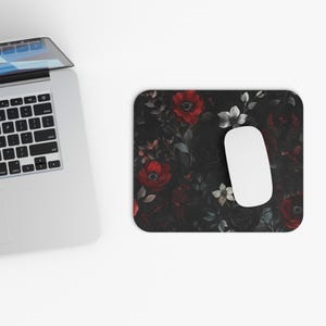 May include: A rectangular mousepad with a floral design featuring red and white flowers on a dark background. A white computer mouse is positioned on the right side of the mousepad. A laptop is visible in the upper left corner.