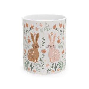 May include: White ceramic mug with a floral pattern featuring two bunnies, one brown and one pink, with white spots. The bunnies are surrounded by green leaves and pink flowers.