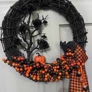 Pode incluir: Uma guirlanda circular de galhos pretos com glitter decorada com rosas pretas, uma pequena abóbora laranja e a silhueta de um gato preto. A base é adornada com cachos de bagas laranjas e pretas, finalizada com uma fita xadrez pied-de-poule.