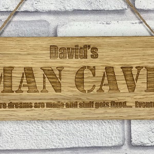 May include: A rectangular wooden sign with rounded corners, featuring the engraved text "David's MAN CAVE". The sign also includes the phrase "Where dreams are made and stuff gets fixed... Eventually." It is made of natural wood and has a rope for hanging.