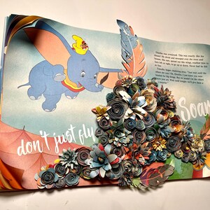 May include: A vintage children's book with a pop-up of a colorful paper flower arrangement. The book features a cartoon image of Dumbo the elephant flying with the text "don't just fly soar" on the page.
