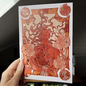 Peut inclure: Une impression d'art représentant une figure aux cheveux roux flottants, tenant un instrument à cordes. L'œuvre utilise des tons chauds d'orange et d'or, avec des motifs floraux et célestes. L'impression est tenue par une main.