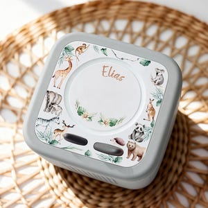 May include: A gray, square-shaped baby monitor with a white top featuring a circular design. The design includes illustrations of various animals like a giraffe, koala, and panda, with the name "Elias" written in script.